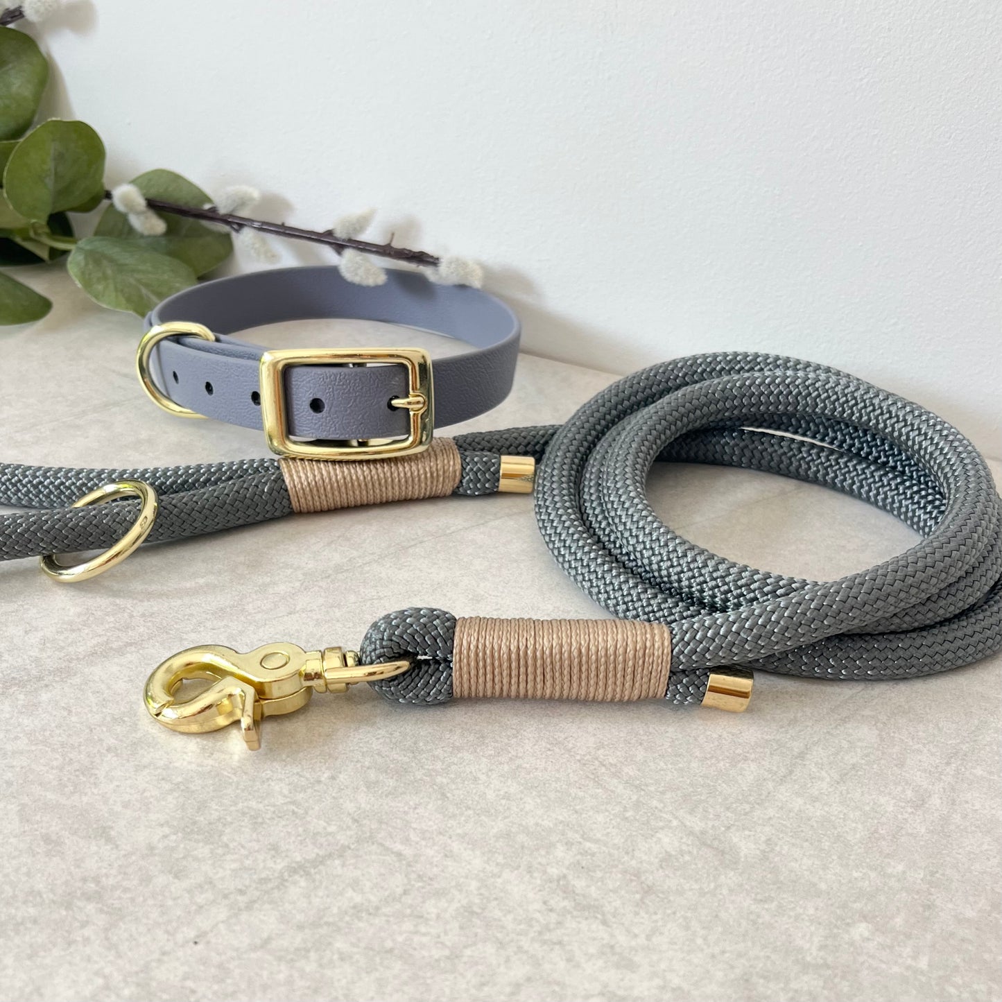 Charcoal Grey rope lead with Light Gold whipping securing a swivel clip at one end and fastening a handle at the other. The handle has an O-ring attached. Each end of the rope is finished with a metal end cap. All hardware for this lead is in Gold. A grey Biothane collar with gold hardware