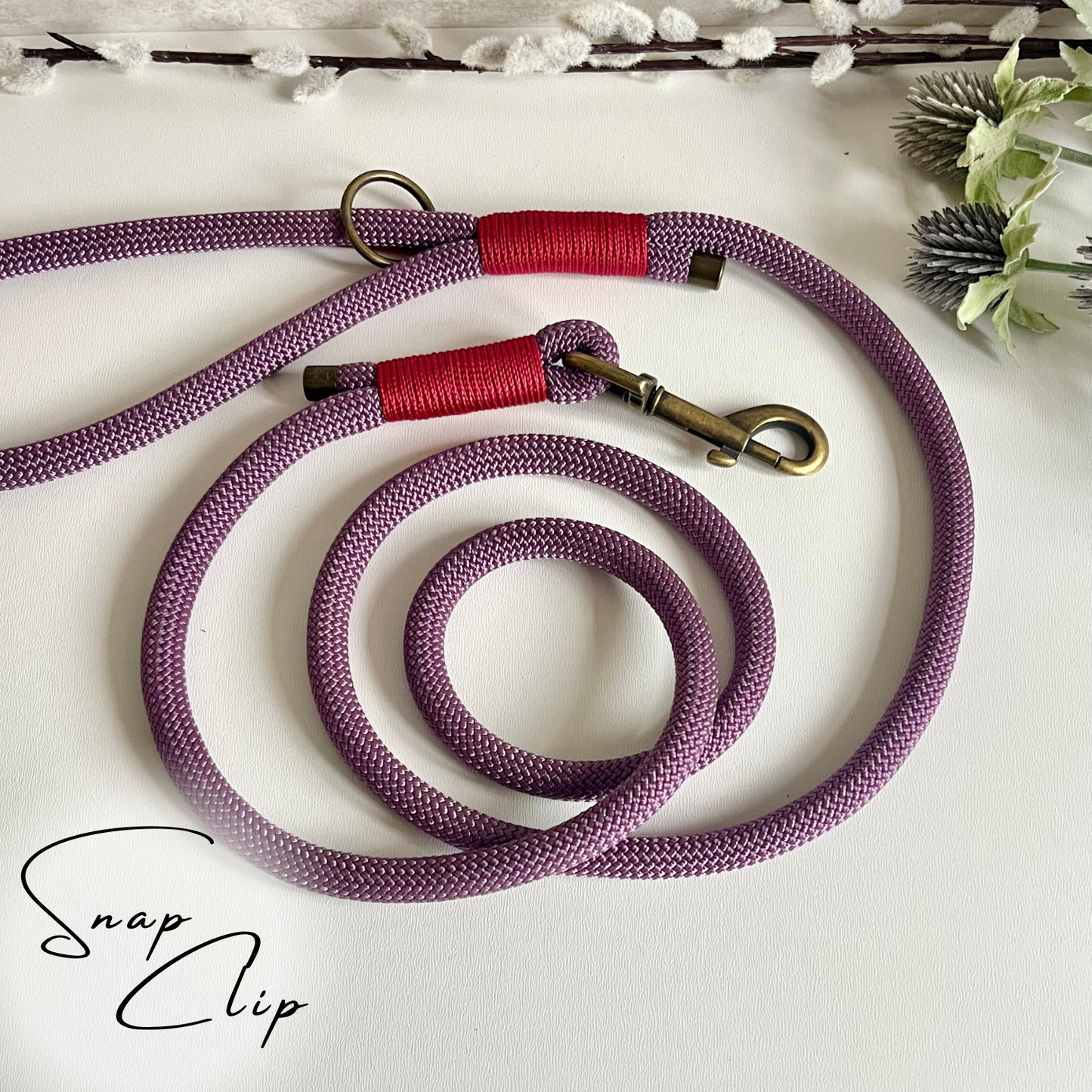 A mauve coloured rope dog lead with raspberry coloured binding and antique brass hardware sitting in a white surface with leaves in the background