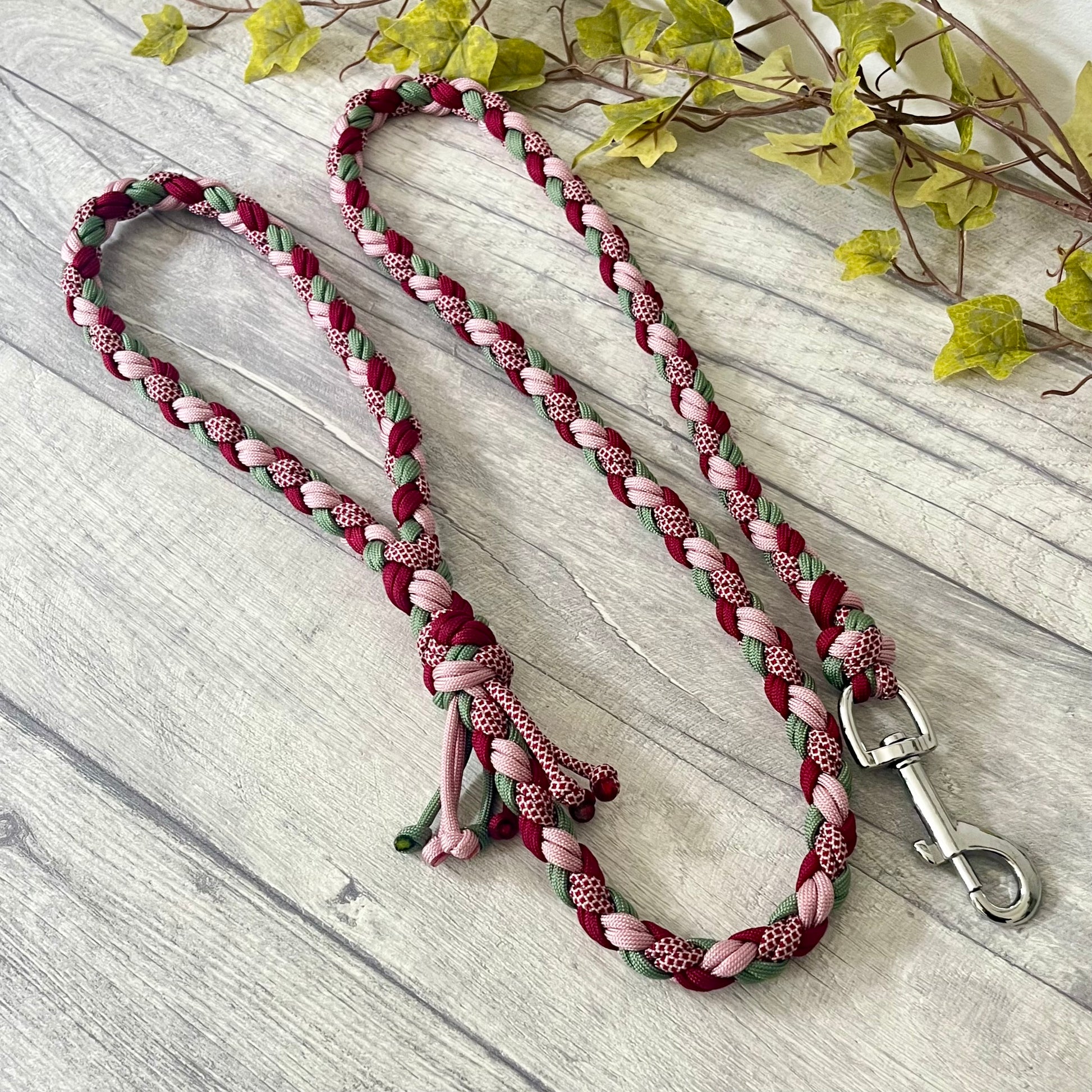 Braided dog leash with pink, green, and red pattern with a silver snap clip on a wooden surface with leaves in the background
