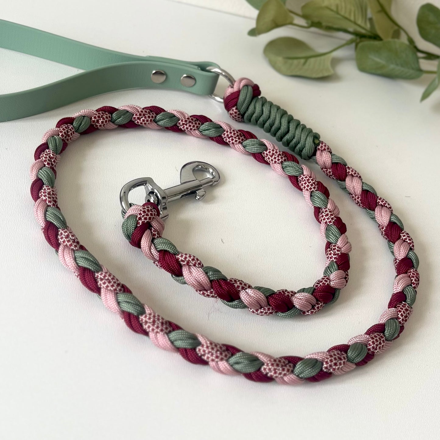 Braided dog leash in pinks, reds and green with green biothane handle white surface with green leaves.