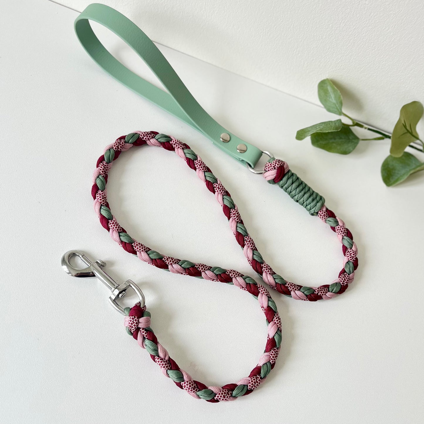 Braided dog leash in pinks, reds and green with green biothane handle white surface with green leaves.