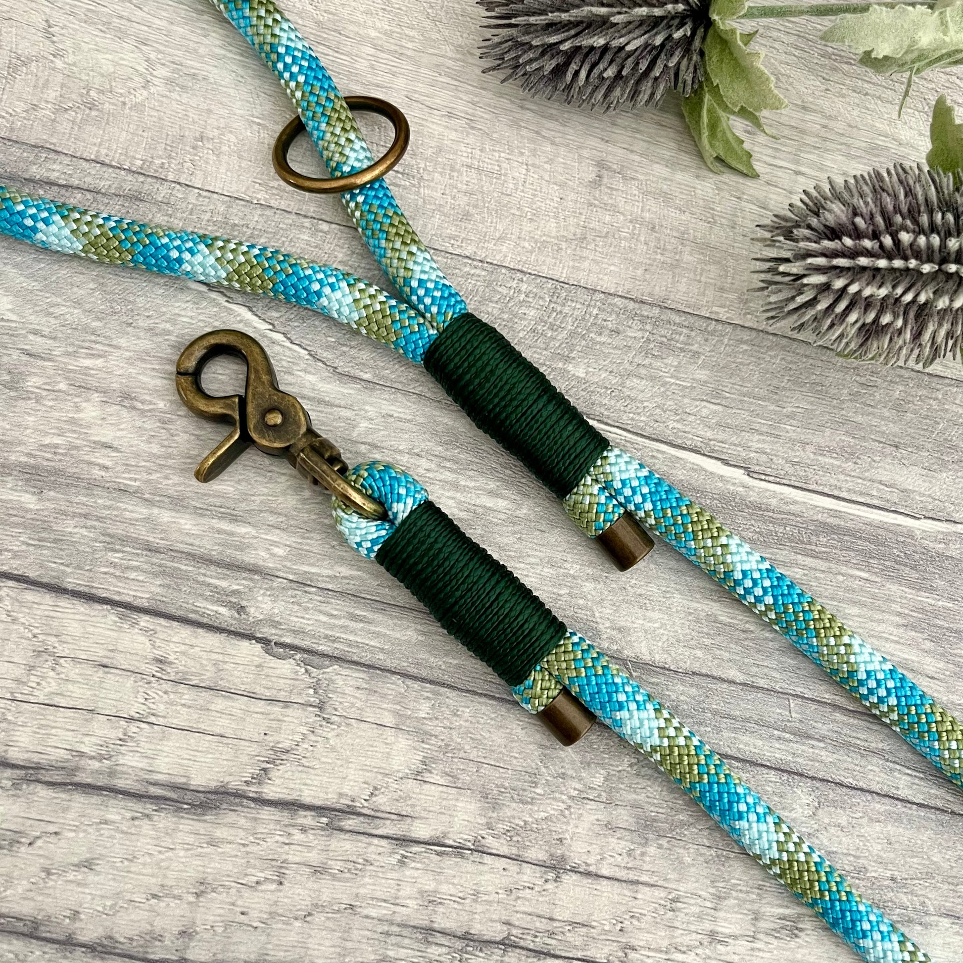 Bombay Blue Paracord rope lead with Dark Green whipping securing each end together. At each end of the rope there is a metal end cap, a clip to connect to a collar at one end and a O-ring fixed to the handle. All hardware  Antique Brass