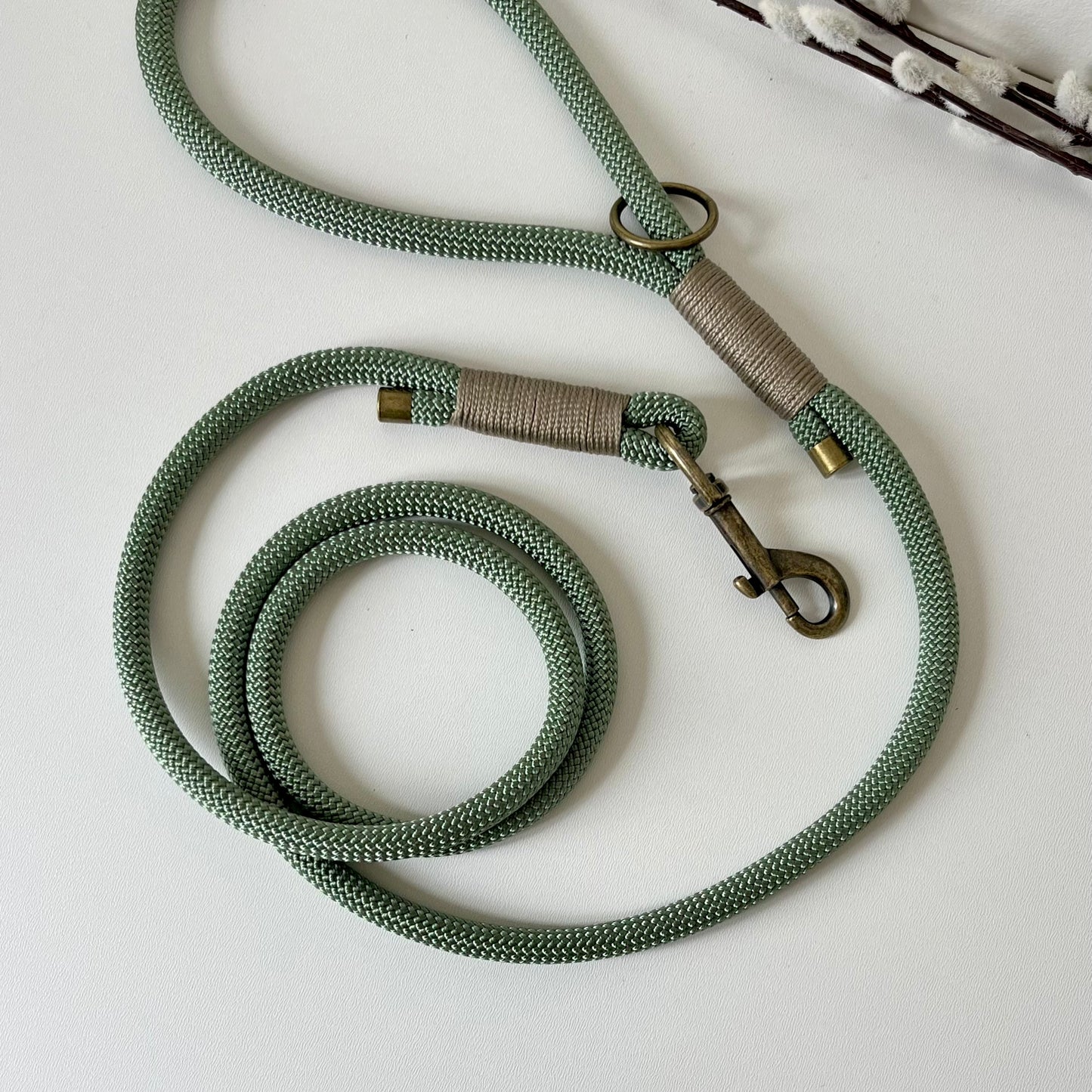 A green rope dog lead with tan binding and antique brass hardware sitting on a white surface