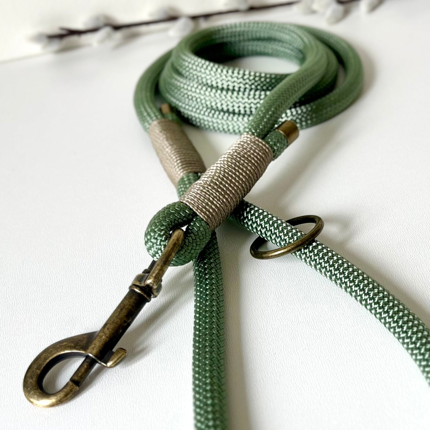 A green rope dog lead with tan binding and antique brass hardware sitting on a white surface