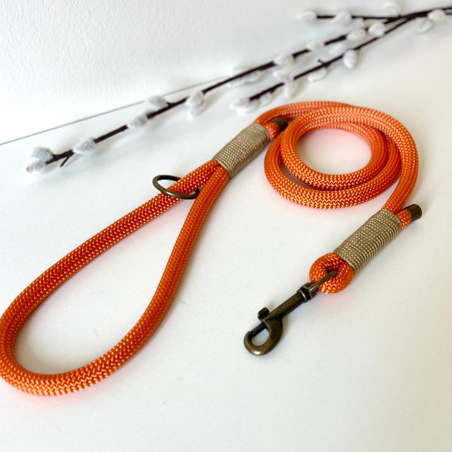 orange rope dog lead with binding and antique brass hardware sitting on a white surface with twigs in the background