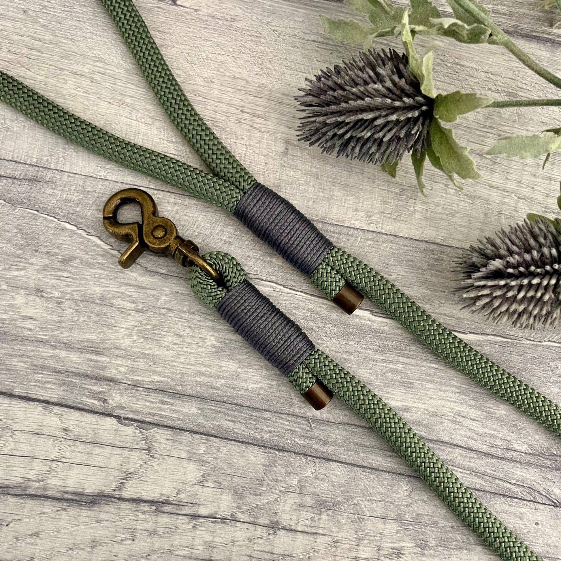 Foggy Green Paracord rope lead with Charcoal whipping securing each end together. At each end of the rope there is a metal end cap, a clip to connect to a collar at one end and a O-ring fixed to the handle. All hardware is Antique Brass