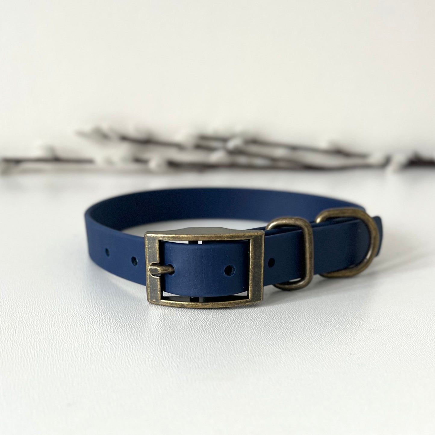 A navy blue Biothane dog collar with antique bronze hardware, displayed on a light gray textured surface with blurred decorative branches in the background.
