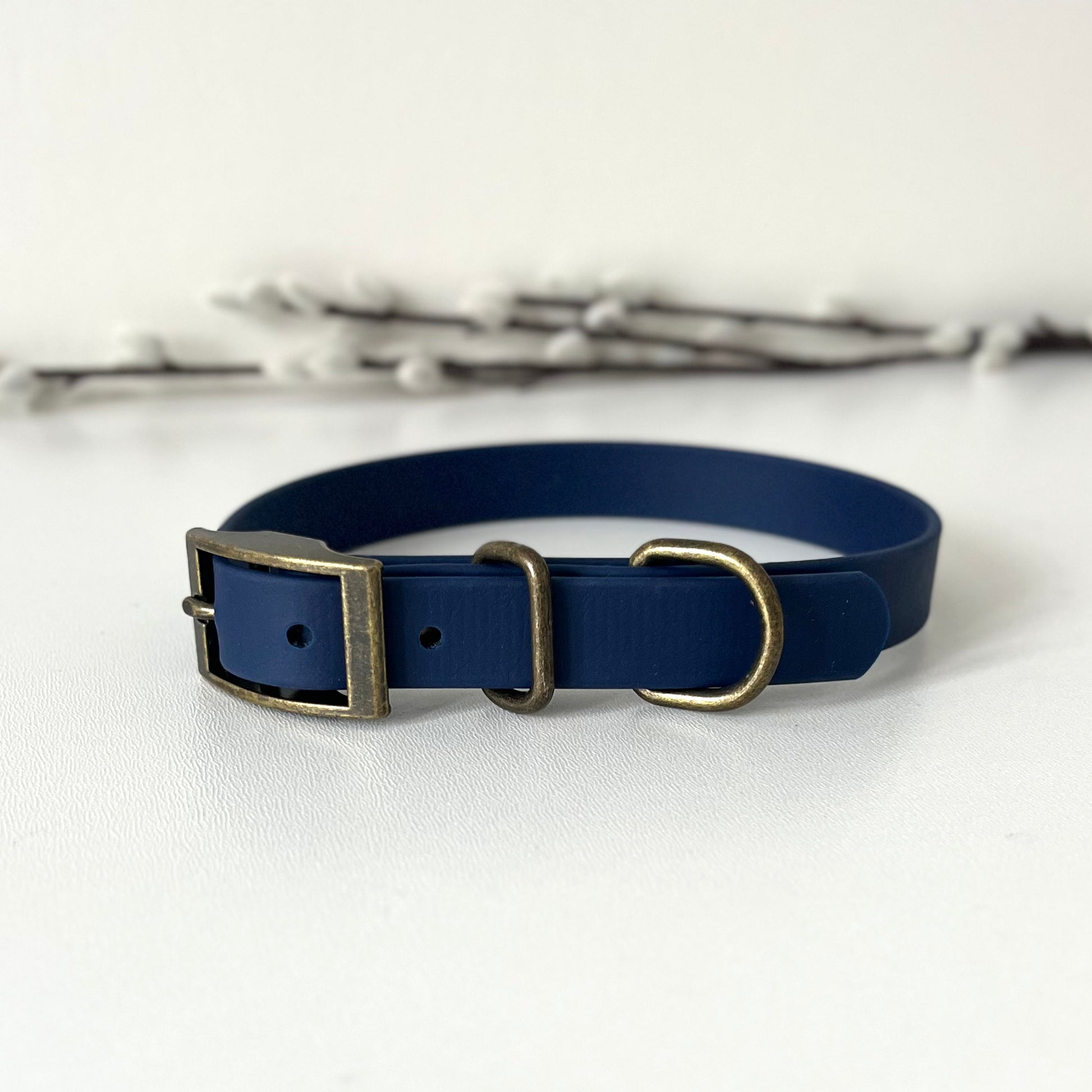 A navy blue Bioithane dog collar with antique bronze hardware, displayed on a white surface with soft-focus white blossoms in the background.