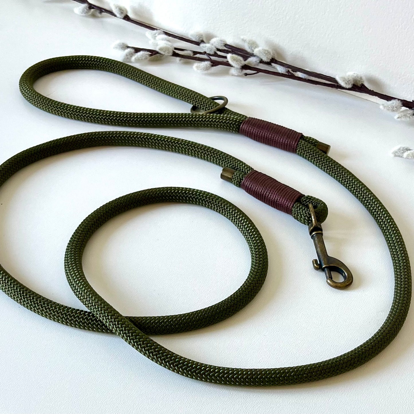 An olive green rope dog lead with brown binding and antique brass hardware sitting on a white surface