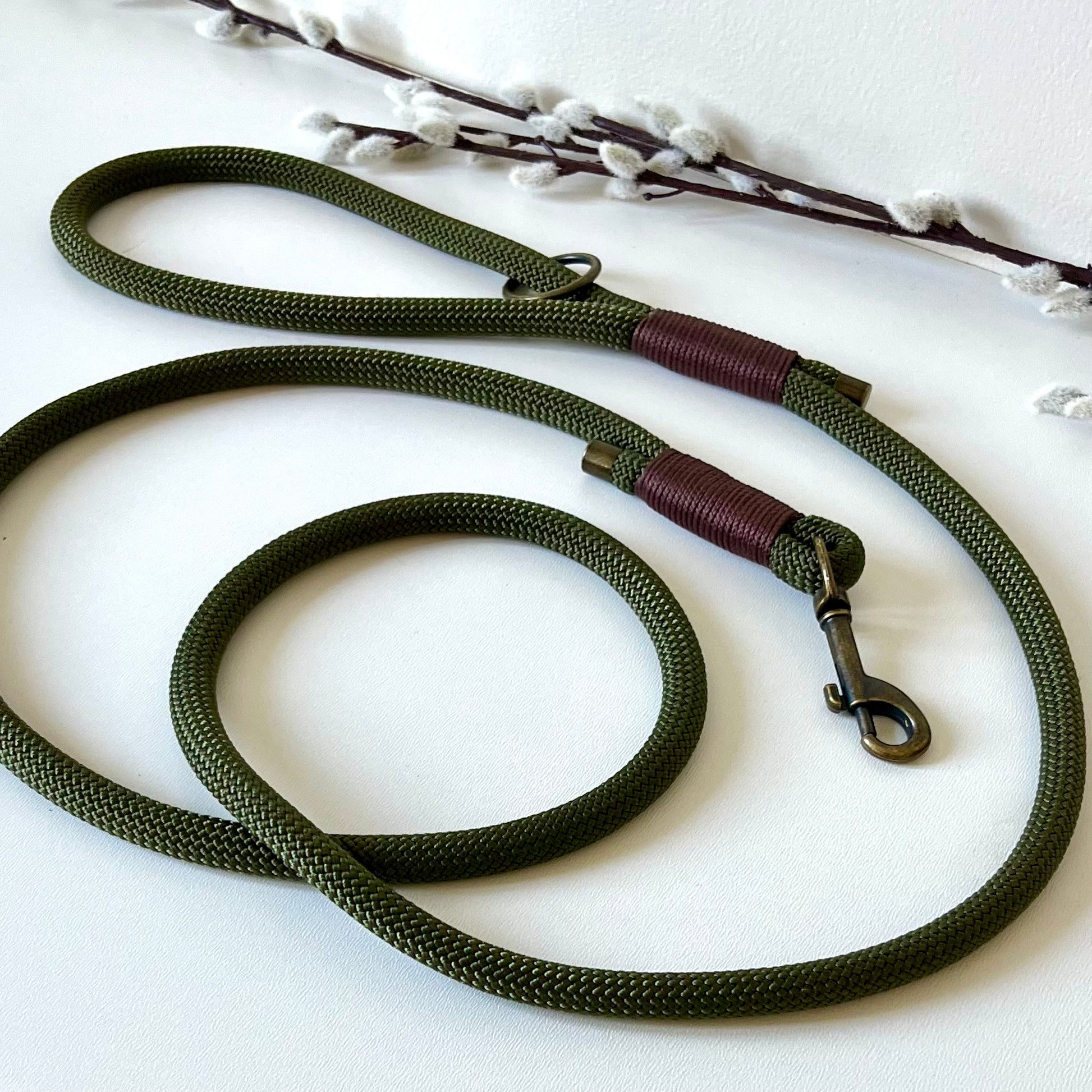 An olive green rope dog lead with brown binding and antique brass hardware sitting on a white surface