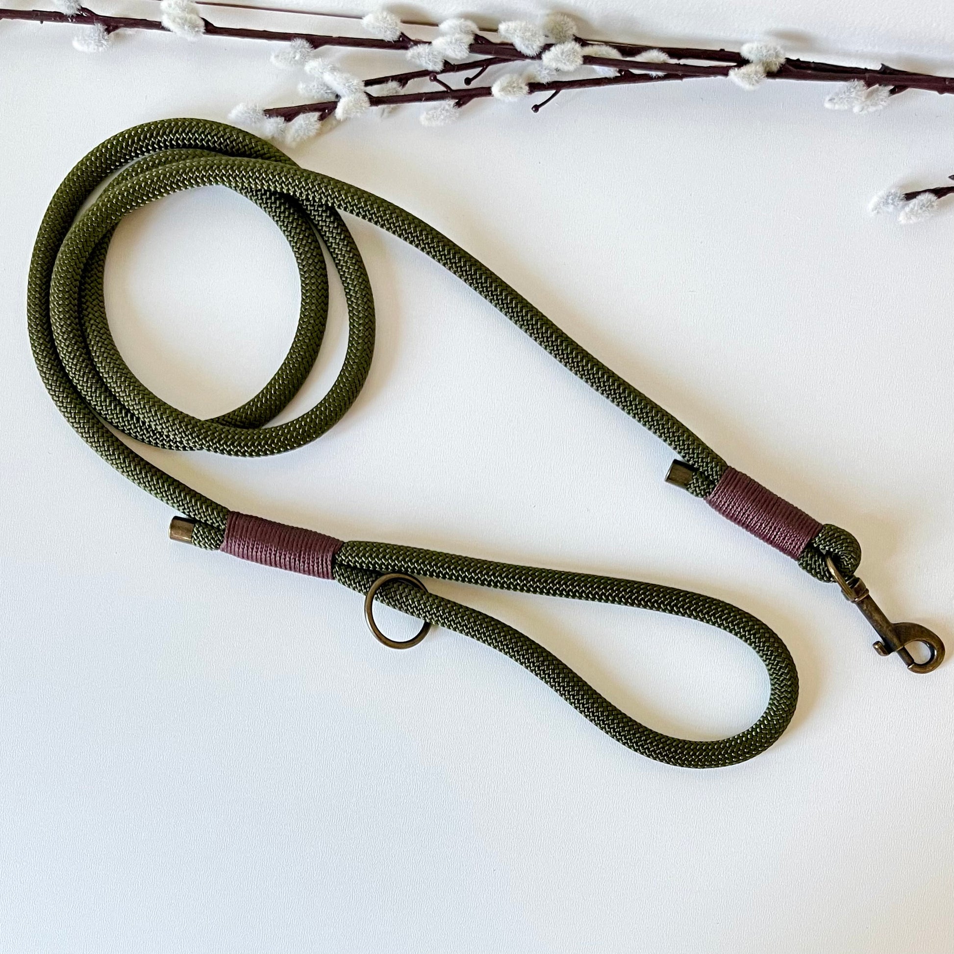 An olive green rope dog lead with brown binding and antique brass hardware sitting on a white surface