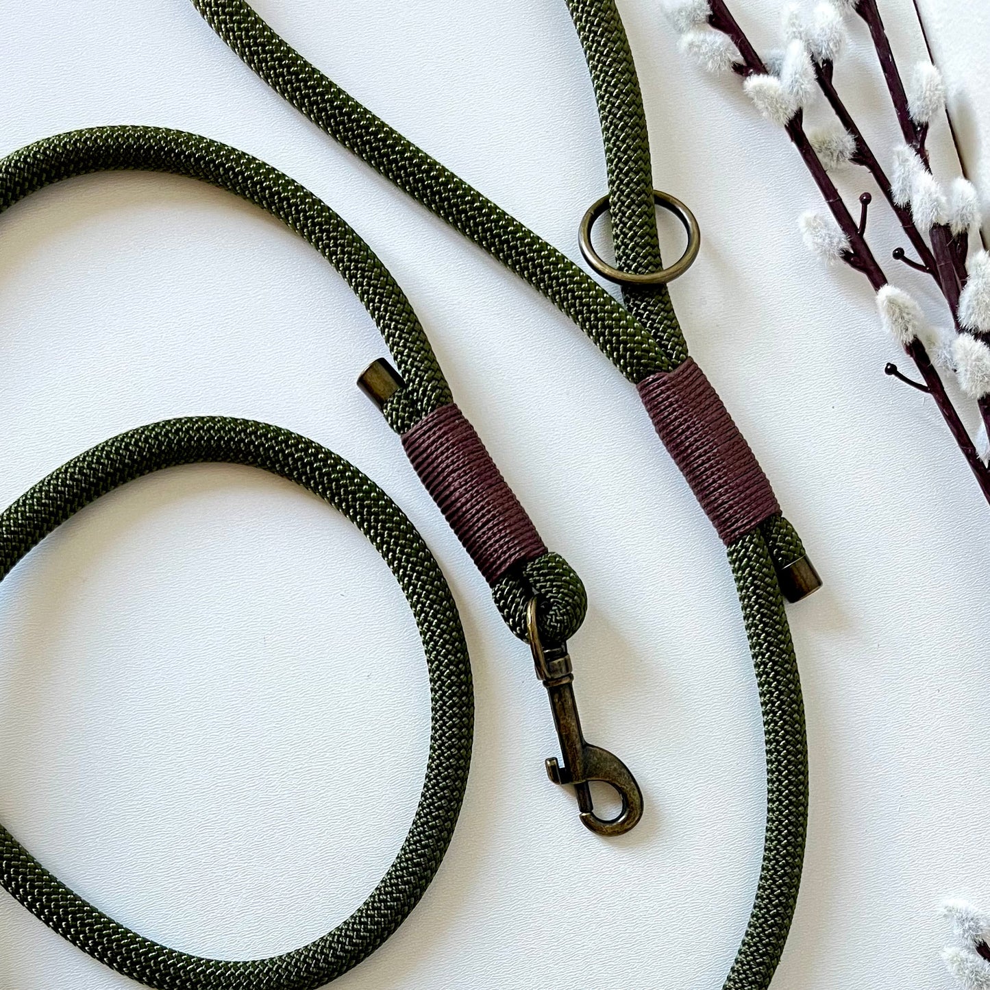 An olive green rope dog lead with brown binding and antique brass hardware sitting on a white surface