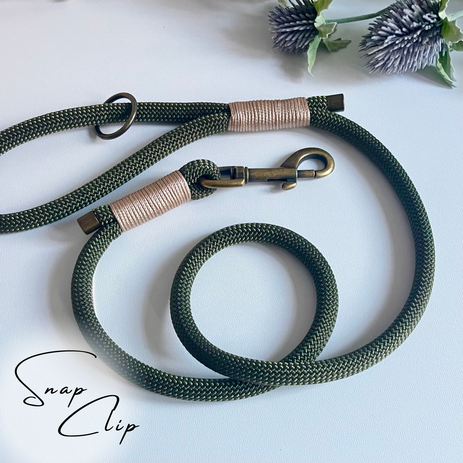 An olive coloured rope dog lead with gold coloured binding and antique brass hardware sitting in a white surface with leaves in the background