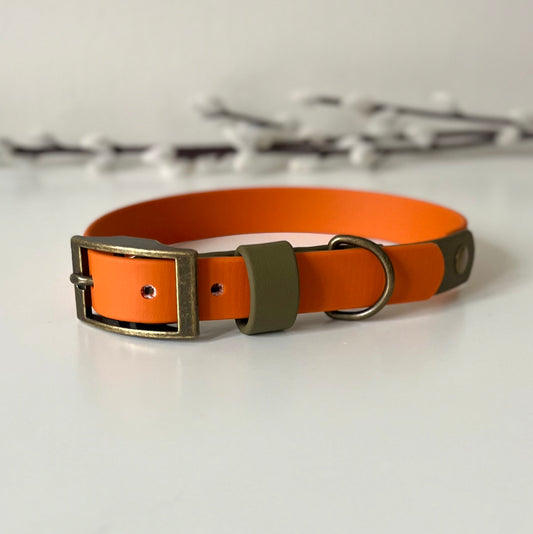 An orange Biothane dog collar with an olive green keeper and antique bronze metal hardware, displayed on a light surface.