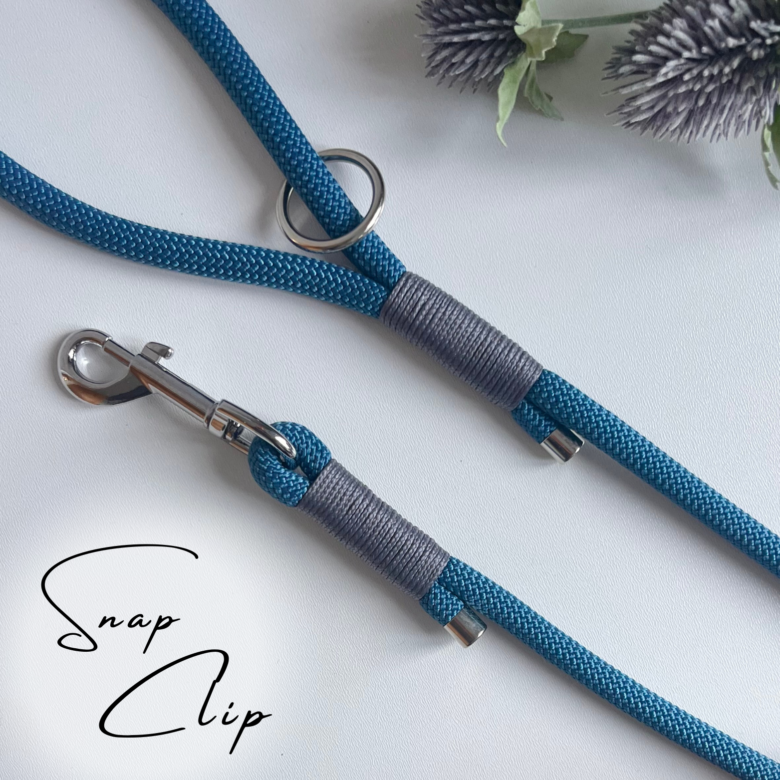 A petrol blue coloured rope dog lead with grey coloured binding and silver hardware sitting in a white surface with leaves in the background