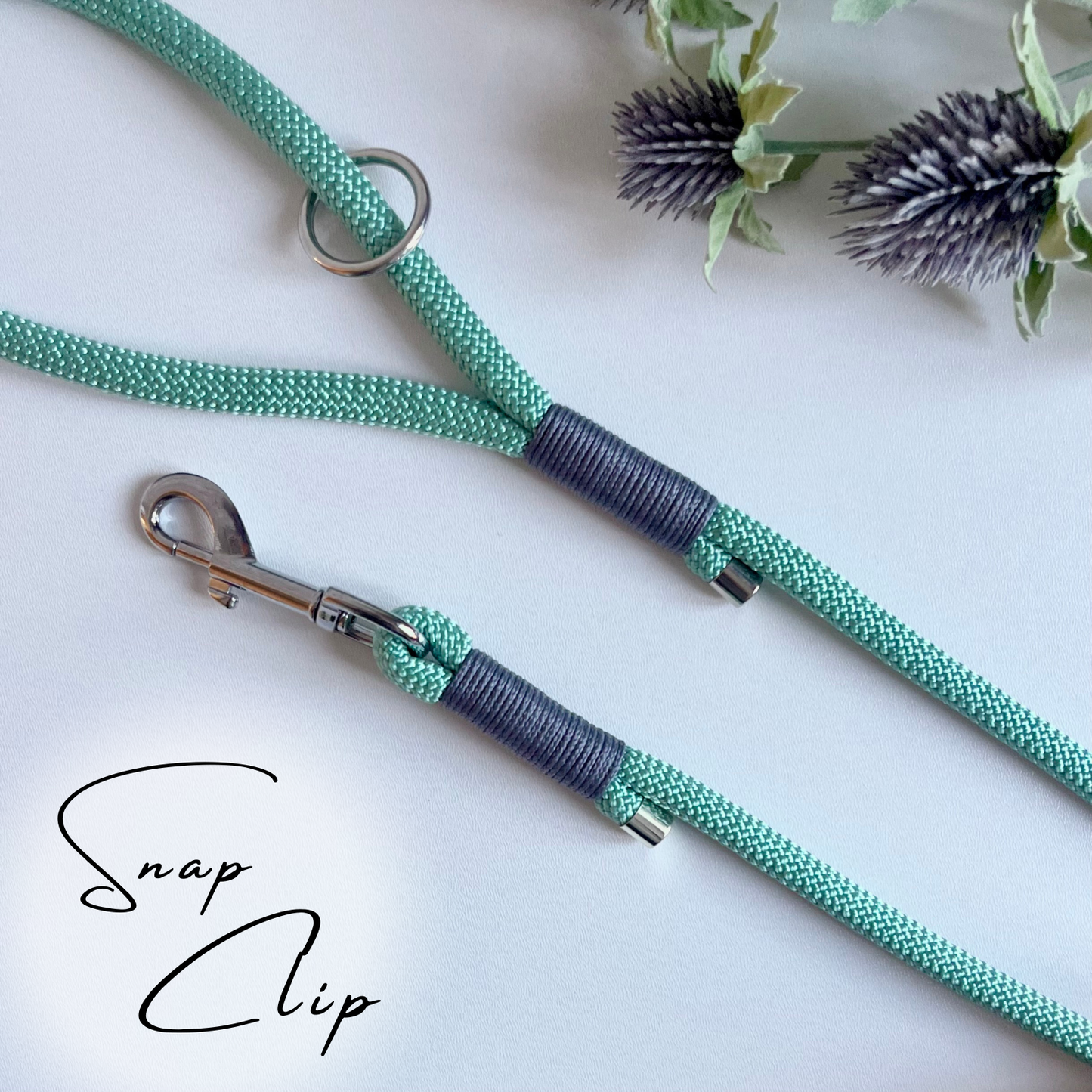 A sea green coloured rope dog lead with grey coloured binding and silver hardware sitting in a white surface with leaves in the background